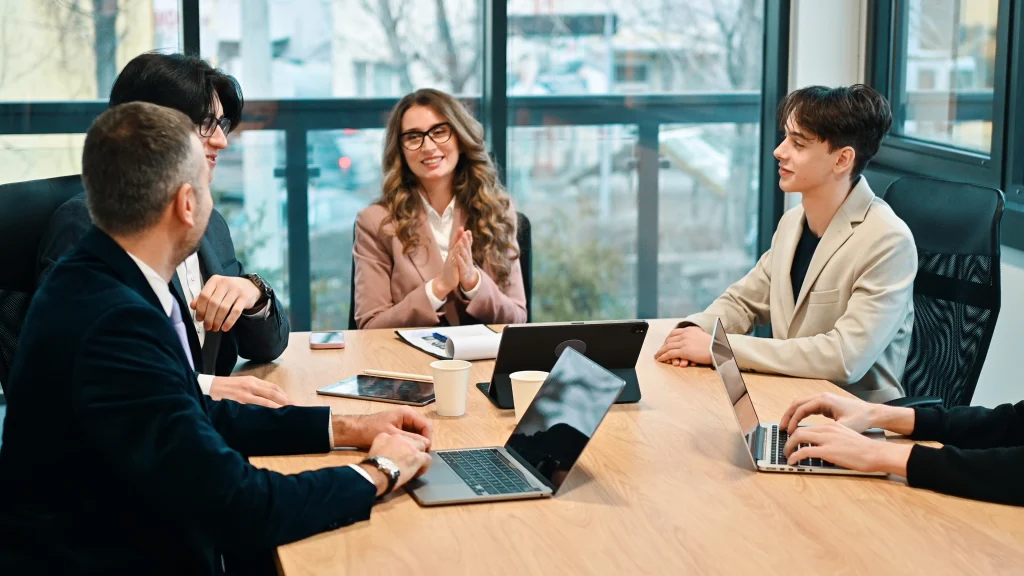 A group of five people sitting around a conference table in a modern office, engaged in a business meeting with laptops, tablets, and documents.
