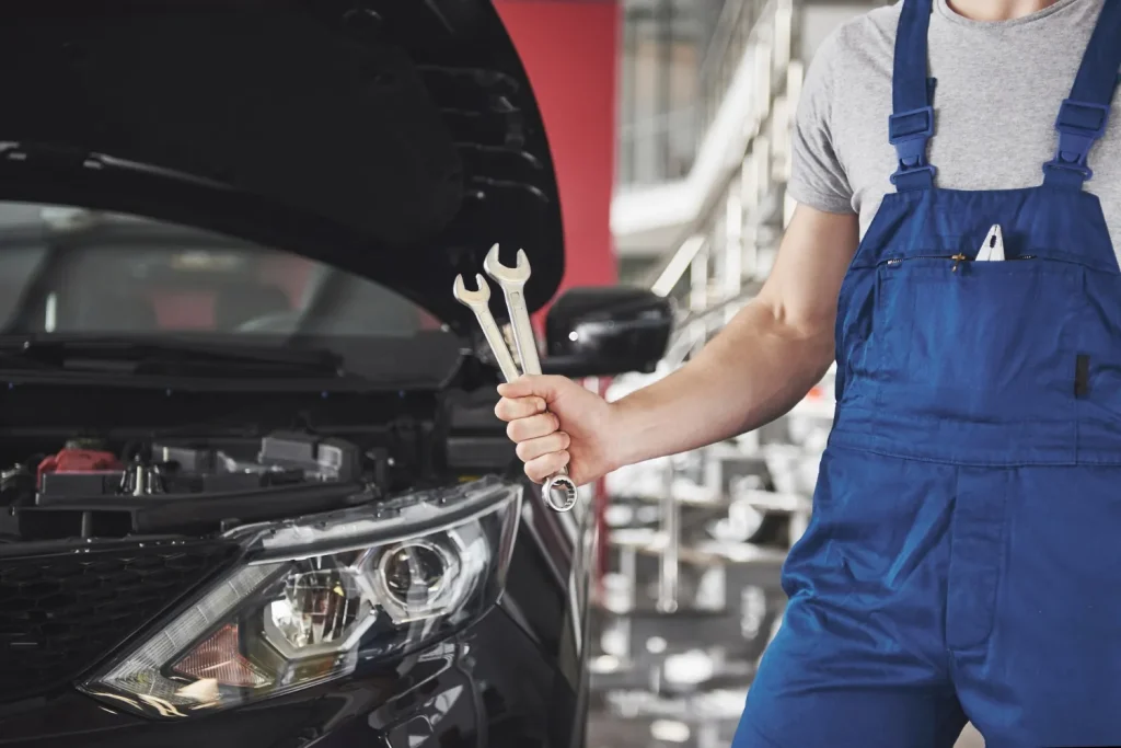 Close-up of a mechanic in blue overalls holding two open-end wrenches in front of the open hood of a black car.