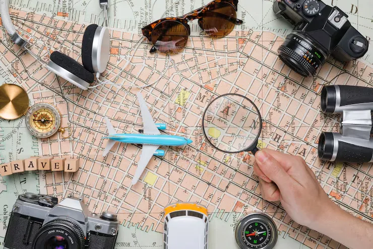 Overhead view of a detailed map surrounded by travel gear, including a toy airplane, sunglasses, headphones, cameras, binoculars, and a magnifying glass.