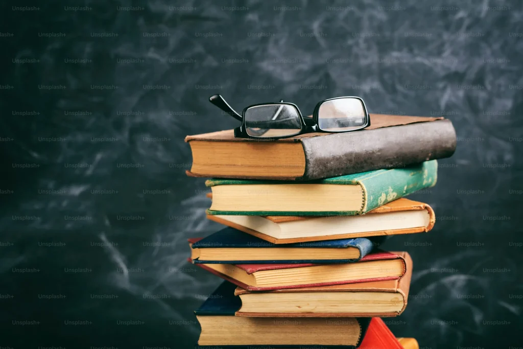 Stack of six old books with a pair of eyeglasses on top, against a dark chalkboard.