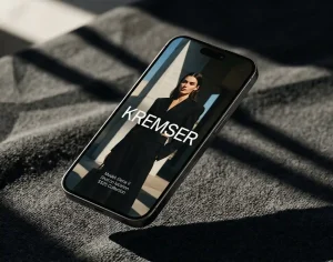 A cinematic, high-angle shot of a smartphone floating diagonally over a dark, textured charcoal fabric. Harsh sunlight creates sharp geometric shadows across the scene. The phone screen displays a high-fashion editorial photo of a model in a black suit with the "KREMSER" logo in bold white typography.