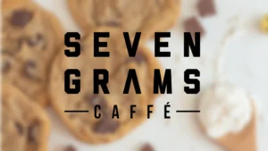 Bold black text reading “SEVEN GRAMS CAFFE” appears over a blurred background of chocolate chip cookies and scattered chocolate pieces on a light surface.