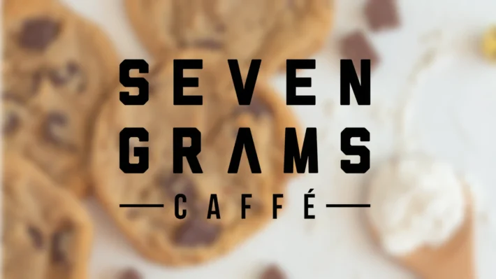 Bold black text reading “SEVEN GRAMS CAFFE” appears over a blurred background of chocolate chip cookies and scattered chocolate pieces on a light surface.