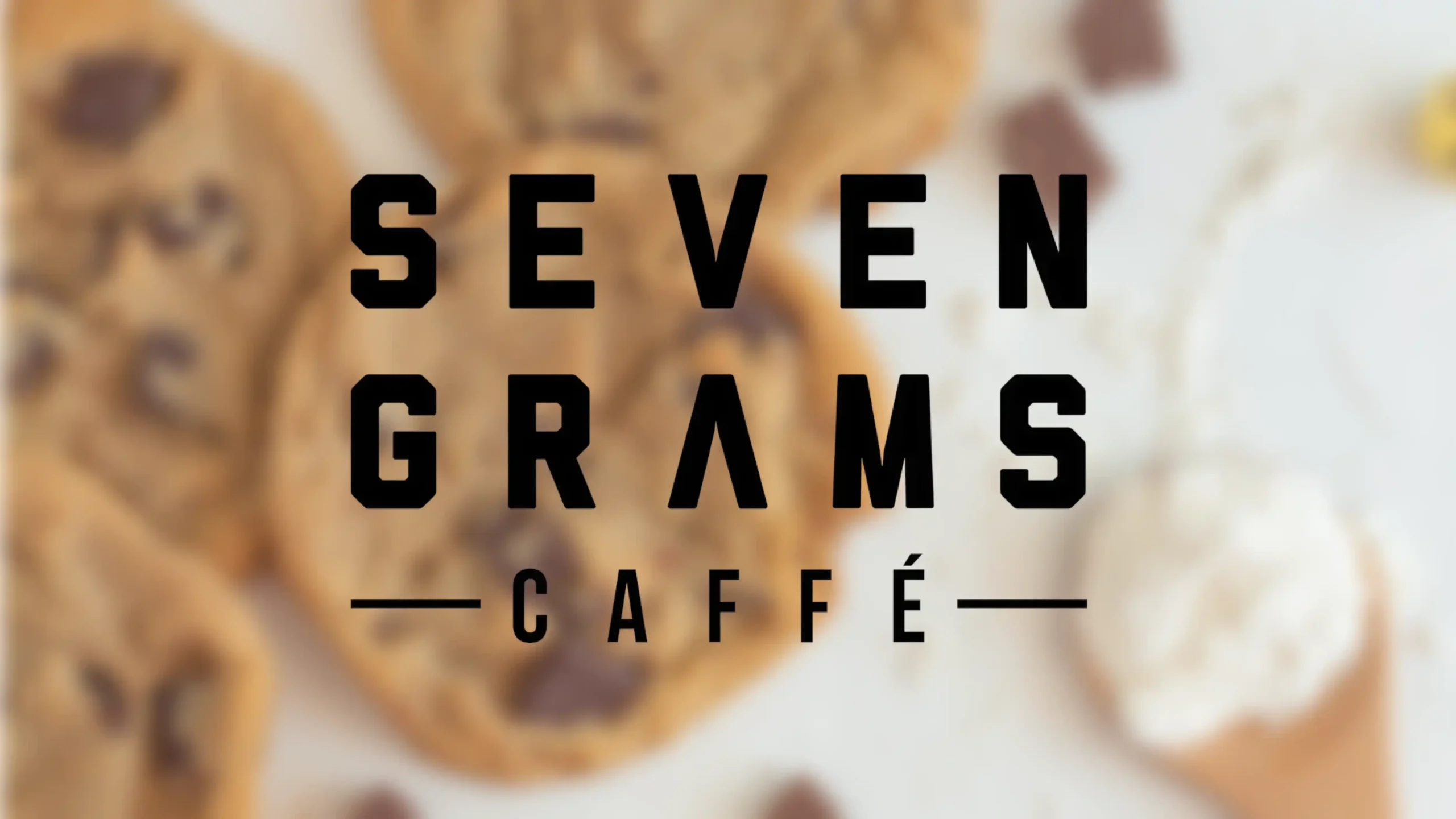 Bold black text reading “SEVEN GRAMS CAFFE” appears over a blurred background of chocolate chip cookies and scattered chocolate pieces on a light surface.