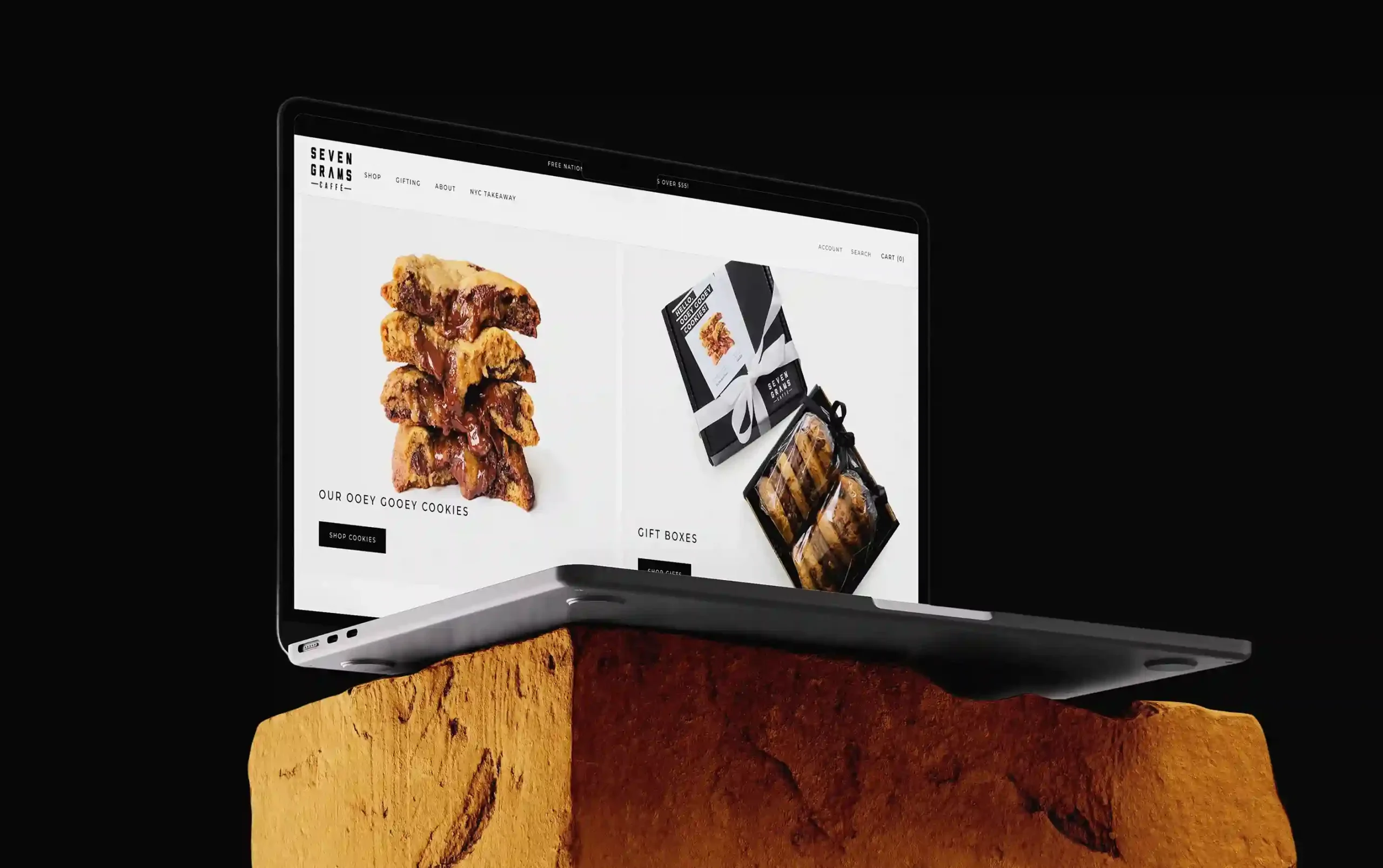 A slim laptop sits partially open on top of a rough stone block against a black background. The screen displays a modern bakery-style e-commerce website featuring stacked chocolate chip cookies with melted filling and packaged gift boxes.