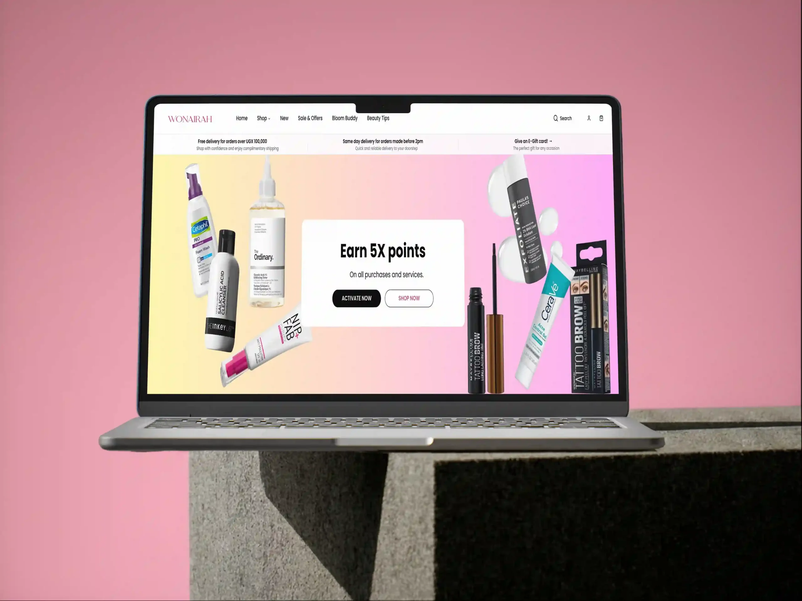 A laptop displayed on a concrete pedestal against a pink gradient background, showing a cosmetics e-commerce website (“WONAIRAH”) with skincare and makeup products floating around the screen and a central promotion banner reading “Earn 5X points” with shop/activate buttons.